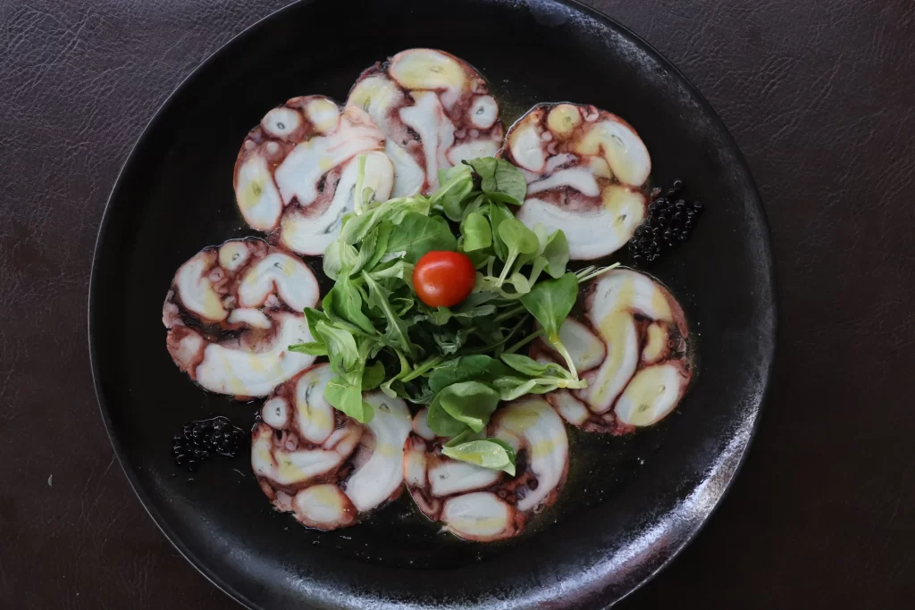Octopus carpaccio arranged in a circular pattern with lamb’s lettuce and cherry tomato garnish at Konoba Malo Misto in Zadar